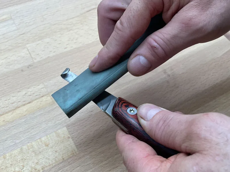 Der Hufschmied schärft ein Hufmesser mit Abziehstein auf Holztisch.