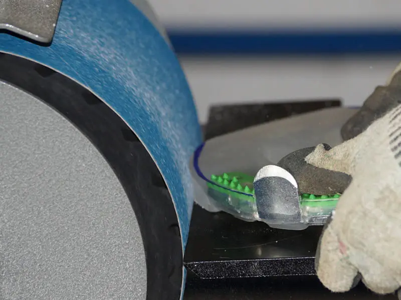 Anpassen eines Hufeisens mit Metallkern und stossdämpfendem Kunststoffmantel am Bandschleifer. Das Hufeisen hat im Zehenbereich eine zusätzliche Stahlkante. Der Hufschmied trägt Schnittschutzhandschuhe.