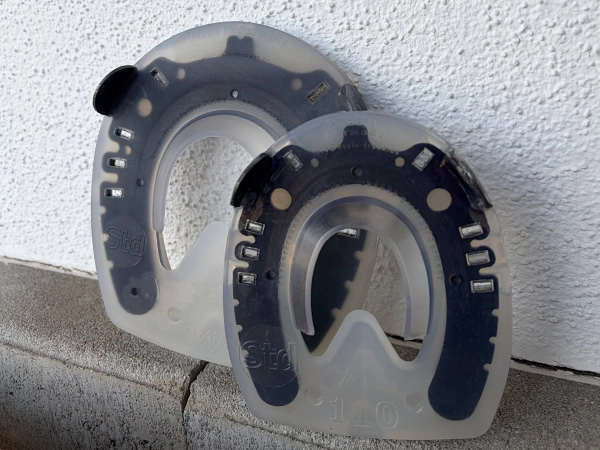 Ein Full Branch Shoe mit regulärem Hufeisenschenkel für mehr Stabilität im Trachtenbereich in der Hufgrösse 110mm steht vor einem Modell in der Grösse 146mm an einer Stallwand.