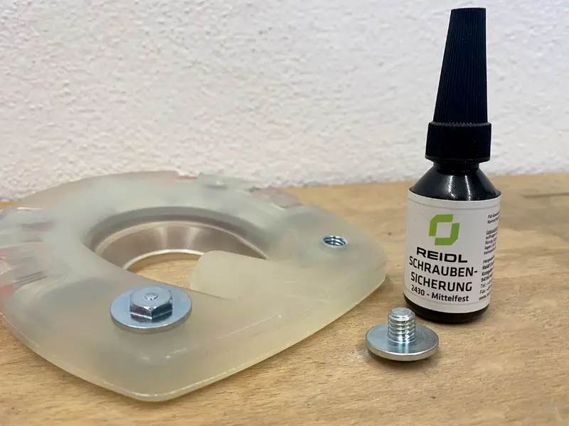 Un ferro convenzionale alternativo con due fori per ramponi nel rivestimento in materiale plastico si trova su un tavolo di legno. Un rampone è già avvitato, un altro è appoggiato davanti. Accanto si trova una bottiglietta di frenafiletti.