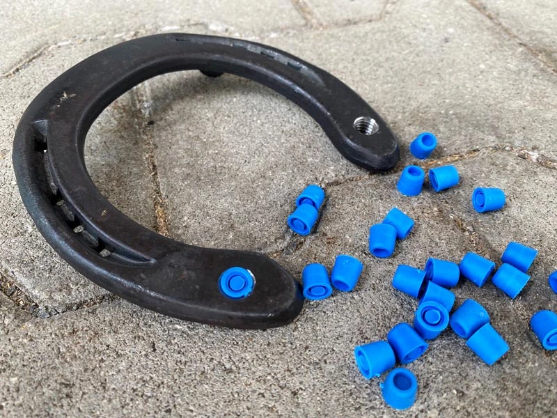 Un fer à cheval repose sur un sol en pierre,
il possède deux trous pour crampons.
L'un des trous est protégé par un bouchon de fermeture bleu.
À côté du fer à cheval se trouvent d'autres bouchons de fermeture en 3/8 pouce.