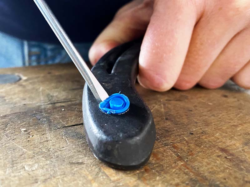 une main tient un fer à cheval, un tournevis plat s'enfonce dans la mortaise à crampon pour retirer le bouchon bleu