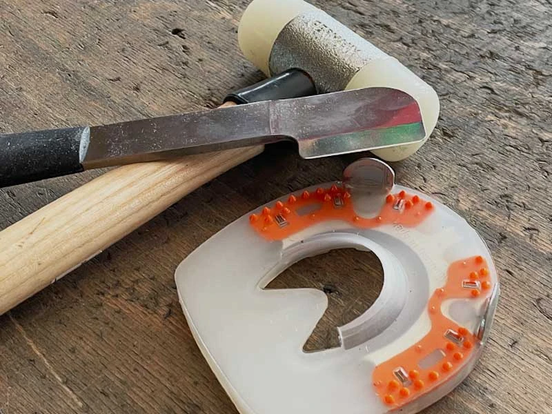 ein Schonhammer mit Nylonkopf, eine Hauklinge in Beilform mit schwarzem ergonomischen Griff und ein stossdämpfendes Hufeisen liegen auf einer Werkbank