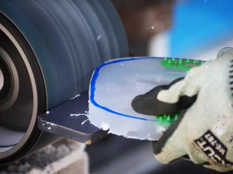 vue sur les mains du maréchal-ferrant ajustant une protection de sabot alternative à la forme du sabot du cheval du client ; une ponceuse à bande apparaît en arrière-plan