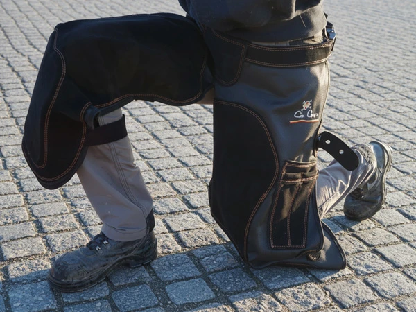 Eine Hufschmiedin trägt das Modell Alex der Schmiedeschürze von Con Cuero. Sie kniet am Boden. Ein dickes Gummipolster im Kniebereich schützt ihr Knie.