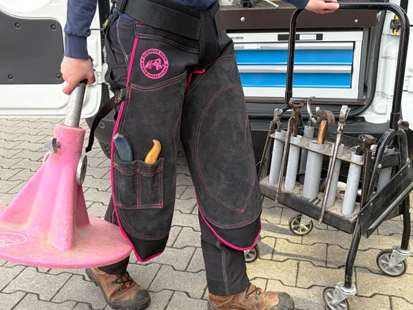 Eine Hufschmiedin trägt die Silverback Chapette Hufschmiedeschürze mit pinken Nähten. In der rechten Hand hält sie einen pinken Hoofjack®, mit der linken Hand fährt sie ihren Werkzeugwagen zum Beschlagsplatz.